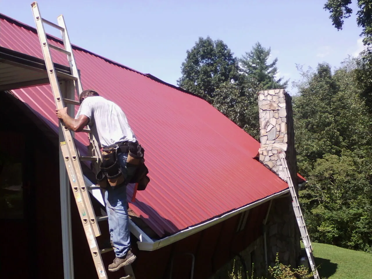 Roofer on a ladder beside a red metal roof and stone chimney — Soffit & Fascia Repair by Roofing Brothers in Garysburg